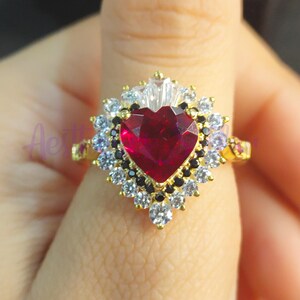 May include: A gold ring featuring a large red heart-shaped gemstone, encircled by smaller white and black gemstones.