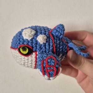 Peut inclure: Peluche Kyogre au crochet faite à la main. Le jouet présente un corps bleu avec des taches blanches, des détails rouges et un ventre blanc. Il a un œil jaune et noir et de petites nageoires. La peluche est tenue sur un fond uni.