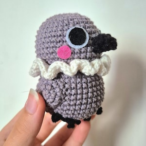 Peut inclure: Un oiseau crocheté à la main, de couleur grise, avec un col blanc à volants, un bec et des pieds noirs. L'oiseau a une joue rose et de grands yeux noirs et blancs. Cet amigurumi fantaisiste est un article unique, fait à la main.