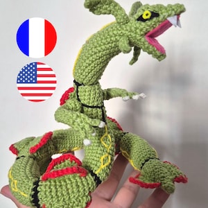 Könnte beinhalten: Eine gehäkelte, drachenähnliche Figur in Grün mit roten Flossen, schwarzen Bändern und gelben Akzenten. Das Objekt hat ein geöffnetes Maul mit weißen Zähnen, gelbe Augen mit schwarzen Pupillen und einen langen, gewundenen Körper mit Krallen.
