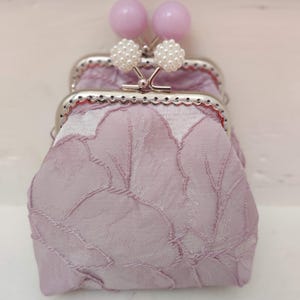 Op de afbeelding: Een kleine, lavendelkleurige portemonnee met een bloemenpatroon. De portemonnee heeft een zilveren sluiting met twee parel-achtige versieringen en twee lavendelkleurige kralen.