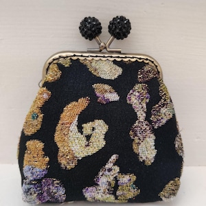 May include: A small black coin purse with a floral pattern in gold, purple, and green. The purse has a metal frame and a clasp with two black rhinestone embellishments.