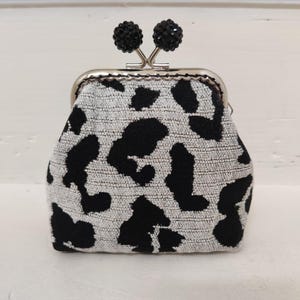 Op de afbeelding: Een kleine portemonnee met een kiss-lock sluiting, zwart en wit van patroon. De portemonnee heeft een zilverkleurig frame en twee zwarte kralenversieringen op de sluiting. De stof heeft een getextureerde uitstraling, die lijkt op een dalmatiërpatroon.