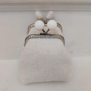 Könnte beinhalten: Eine kleine weiße Filz-Münzbörse mit einem silbernen Metallrahmen und zwei weißen perlmuttartigen Knöpfen.