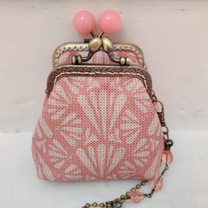 Op de afbeelding: Een kleine roze stoffen portemonnee met een bloemenpatroon en een messing sluiting. De portemonnee heeft een ketting met roze kralen.