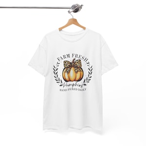 May include: White t-shirt featuring a pumpkin design with a leopard print bow, surrounded by a wreath. The text reads "FARM FRESH PUMPKINS HAND PICKED DAILY". The shirt is hanging on a wooden hanger.