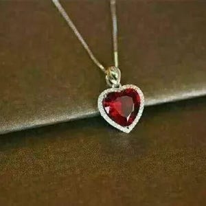 May include: A silver heart-shaped pendant necklace with a large red gemstone in the center. The gemstone is surrounded by a row of smaller white gemstones.