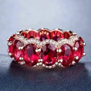 May include: A red ruby and diamond eternity band ring in rose gold. The ring features a row of oval-shaped rubies set in a pave setting with smaller diamonds surrounding each ruby.
