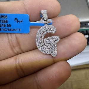May include: A silver pendant featuring a large letter G crafted from pave set diamonds. The pendant is on a chain and is being held by a hand.