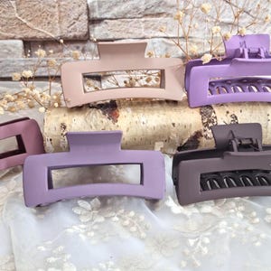 Könnte beinhalten: Fünf matte Haarklammern in den Farbtönen Mauve, Lavendel und Braun. Die Klammern sind rechteckig mit einem ausgeschnittenen Mittelteil und einem Scharnierverschluss. Sie sind auf einem Birkenstamm und weißem Spitzengewebe angeordnet.