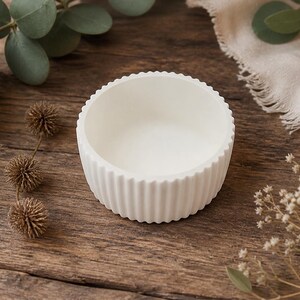 Könnte beinhalten: Eine kleine, weiße, runde Schale mit gerippter Außenseite. Die Schale steht auf einer rustikalen Holzoberfläche, umgeben von Zweigen mit Grün und Trockenblumen. Das Design der Schale ist schlicht und elegant.