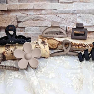 Puede incluir: Una colección de pinzas para el cabello en varias formas y colores. Las pinzas incluyen un diseño retorcido negro, una forma de flor y varias opciones de tonos neutros. El fondo presenta una textura de madera y una tela de encaje blanco.