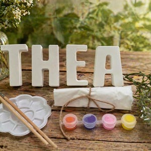 Könnte beinhalten: Ein Bastelset mit dem Namen "THEA" in weißen Keramikbuchstaben. Das Set enthält Farbtöpfe, Pinsel und eine Palette, die auf einer Holzoberfläche angeordnet sind. Eine Vase mit Blumen befindet sich im Hintergrund.