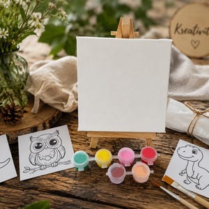 Könnte beinhalten: Eine leere Leinwand auf einer kleinen Holzstaffelei, umgeben von Kunstzubehör. Enthält Farbtöpfe, Pinsel und Malvorlagen mit Dinosaurier- und Eulenillustrationen. Das Wort "Kreativitat" steht auf einer Holzscheibe.