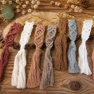 Könnte beinhalten: Eine Auswahl handgefertigter Makramee-Schlüsselanhänger in verschiedenen Farben, darunter Rot, Weiß, Braun, Grau und Rosa. Jeder Schlüsselanhänger hat ein geflochtenes Design und eine lange Quaste, befestigt an einem goldfarbenen Ring. Die Schlüsselanhänger liegen auf einer Holzoberfläche.