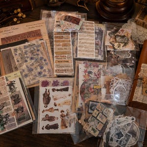 Könnte beinhalten: Eine Sammlung von Scrapbooking-Materialien im Vintage-Stil, bestehend aus verschiedenen Stickerbögen, Papier-Ephemera und dekorativen Ausschnitten mit botanischen, historischen und textbasierten Motiven sowie einem braunen Lederjournal.