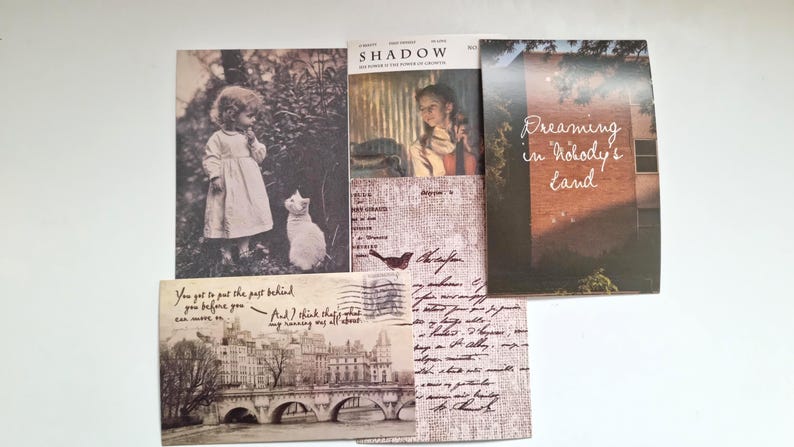 Op de afbeelding: Een verzameling ansichtkaarten in vintage-stijl en een tijdschrift. De ansichtkaarten tonen sepia-kleurige afbeeldingen van een kind met een kat, een brug en handgeschreven tekst. De omslag van het tijdschrift luidt "SHADOW" en "Dreaming in Nobody's Land".