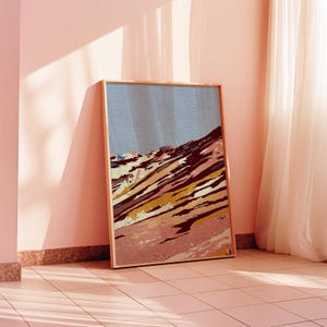 Könnte beinhalten: Ein gerahmter Kunstdruck mit einer abstrakten Berglandschaft. Das Kunstwerk verwendet eine Farbpalette aus Rosa, Braun, Gelb und Blau. Der Rahmen ist helles Holz, und der Druck lehnt an einer rosa Wand.