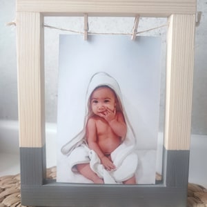 Puede incluir: Un marco de fotos de madera con la parte inferior gris y la superior de madera natural. Una foto de un bebé con una toalla con capucha blanca se muestra en el marco.