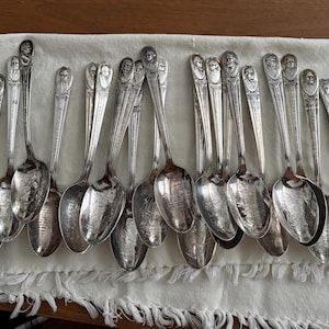 May include: A collection of antique silver spoons arranged on a white fringed cloth. Each spoon features a detailed portrait on the handle and a reflective bowl. The spoons are displayed on a wooden surface, showcasing their intricate design.