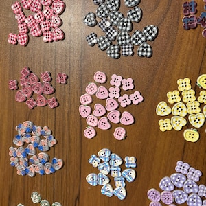 May include: A collection of decorative buttons in various shapes and colors. The buttons include heart, flower, and round shapes, with patterns like gingham and stripes. Colors include pink, blue, purple, yellow, and black and white.