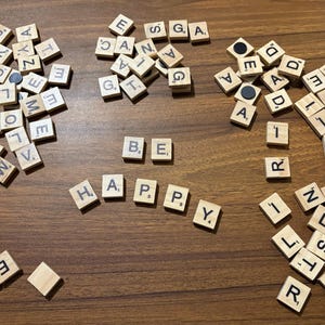 May include: Wooden Scrabble tiles spelling "BE HAPPY" on a brown wooden surface. Scattered tiles with letters are arranged around the phrase. The tiles are light brown with black letters.