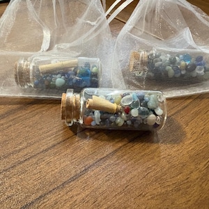 May include: Three small glass bottles filled with colorful gemstones and a rolled-up paper scroll, each sealed with a cork. The bottles are presented in sheer white drawstring bags, set against a wooden surface.
