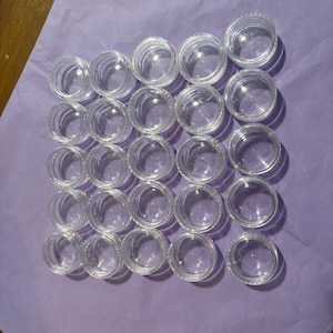 May include: A collection of small, clear plastic containers with screw-on lids, arranged neatly on a purple surface. The containers are round and transparent, allowing visibility of the contents. Ideal for storing small items.