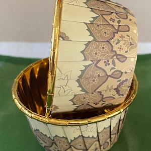 May include: Two decorative cupcake liners. The liners are beige with a floral pattern in shades of brown and gray. The rims and handle are gold. One liner is nested inside the other, creating a visually appealing arrangement.