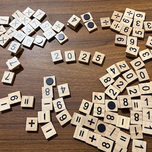 May include: Wooden number tiles and math symbols scattered on a brown surface. The tiles spell out "2025" and include numbers, plus, minus, division, and multiplication symbols. Some tiles have black magnetic dots.