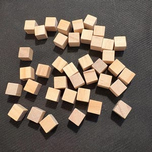 May include: A collection of small, unfinished wooden cubes scattered on a dark surface. The cubes are a light, natural wood color, and each is approximately 0.75 inches square. The cubes appear to be game pieces or craft supplies.