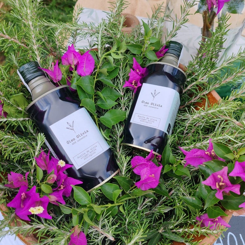 Puede incluir: Dos botellas oscuras de aceite de oliva con etiquetas blancas, rodeadas de vibrantes flores de buganvillas rosas y follaje verde. Las etiquetas dicen "&Oacute;leo Olivia" y "Aceite de oliva virgen extra ecol&oacute;gico."