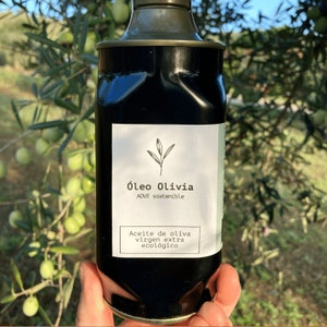 Puede incluir: Una botella oscura de aceite de oliva con una etiqueta blanca que muestra el texto "&Oacute;leo Olivia" y "Aceite de oliva virgen extra ecol&oacute;gico." La botella tiene una tapa met&aacute;lica. El fondo muestra ramas de olivo y aceitunas verdes.