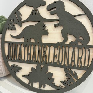 May include: A round wooden sign featuring a green dinosaur silhouette and the names "Jonathan & Leonard" in a black font.