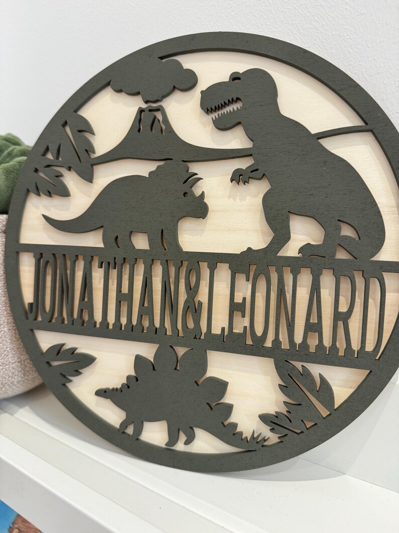May include: A round wooden sign featuring a green dinosaur silhouette and the names "Jonathan & Leonard" in a black font.