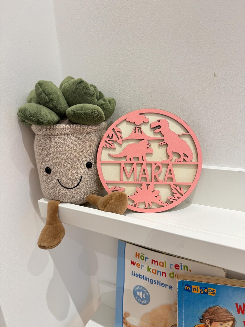 May include: Pink wooden dinosaur-themed name sign with the name "MARA" cut out in the centre. The sign is on a white shelf with a plush succulent plant toy.