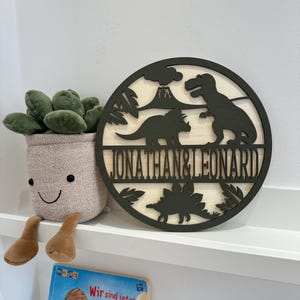 May include: A round wooden sign featuring a black silhouette of dinosaurs and a volcano. The sign is painted green and bears the names "Jonathan & Leonard" inscribed on it.