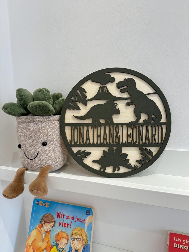 May include: A round wooden sign featuring a black silhouette of dinosaurs and a volcano. The sign is painted green and bears the names "Jonathan & Leonard" inscribed on it.