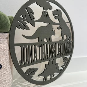May include: A round wooden sign with a dinosaur theme. The sign features a volcano, palm trees, and three dinosaurs. The text "JONATHAN & LEONARD" is cut out of the wood.