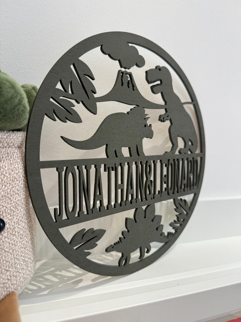 May include: A round wooden sign with a dinosaur theme. The sign features a volcano, palm trees, and three dinosaurs. The text "JONATHAN & LEONARD" is cut out of the wood.
