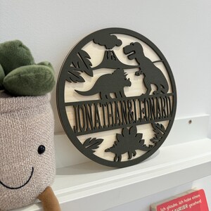 May include: A round wooden sign with a dinosaur theme. The sign is painted black and features a volcano, palm trees, and three dinosaurs. The names "Jonathan & Leonard" are written in a black banner across the bottom of the sign.