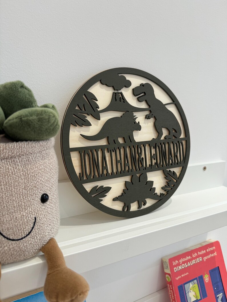May include: A round wooden sign with a dinosaur theme. The sign is painted black and features a volcano, palm trees, and three dinosaurs. The names "Jonathan & Leonard" are written in a black banner across the bottom of the sign.