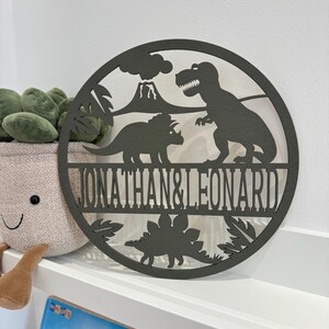 May include: A round wooden sign with a dinosaur theme. The sign features a T-Rex, a Triceratops, and a volcano. The names "Jonathan & Leonard" are written in a decorative font on the sign.