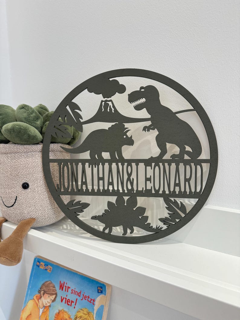 May include: A round wooden sign with a dinosaur theme. The sign features a T-Rex, a Triceratops, and a volcano. The names "Jonathan & Leonard" are written in a decorative font on the sign.