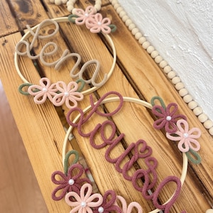 Könnte beinhalten: Zwei florale Namensreifen mit den Namen "Lena" und "Willow" in Schreibschrift. Die Reifen sind mit gestrickten Blumen in Rosa, Grün und Burgunder verziert, mit Perlen in der Mitte. Der Hintergrund umfasst Holzperlen und eine Holzoberfläche.