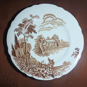 May include: A round, off-white ceramic plate with a scalloped edge, featuring a brown transferware print. The design depicts a rural landscape with a cottage, a body of water, trees, and floral motifs along the bottom and side borders.