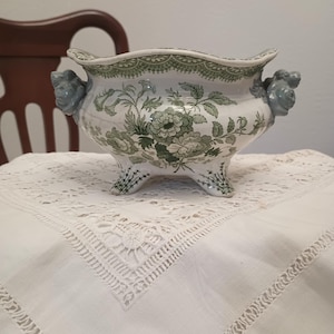 May include: A white ceramic bowl with a scalloped rim and three feet, featuring a green floral pattern and two grey sculpted rose-shaped handles on the sides. The bowl has a decorative green lace-like border along the top edge.
