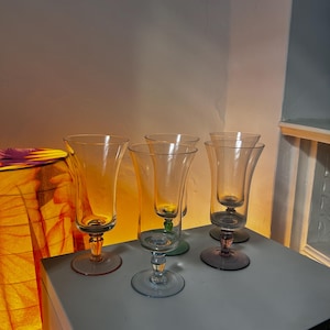 May include: A set of five clear glass goblets with slender stems and circular bases. Each glass features a distinctively coloured base, including shades of amber, blue, green, and purple, supporting a tall, smooth, and transparent bowl.