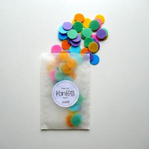 Może przedstawiać: Przezroczysty plastikowy woreczek wypełniony kolorowymi k&oacute;łkami konfetti z filcu w odcieniach niebieskiego, zielonego, ż&oacute;łtego, pomarańczowego i fioletowego. Woreczek ma białą etykietę z napisem "Made with Konfetti regn  pastel".