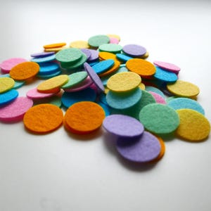 Może przedstawiać: Stosik kolorowych k&oacute;łek z filcu w r&oacute;żnych odcieniach niebieskiego, zielonego, ż&oacute;łtego, pomarańczowego i fioletowego. K&oacute;łka są ułożone w przypadkowy wz&oacute;r i wszystkie są tej samej wielkości.